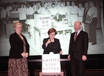 Two women and a man stand in front of a sign that says "SAMMS STOP BUTTON" in front of a historical photo of a group of employees about to hit the "SAMMS GO BUTTON."