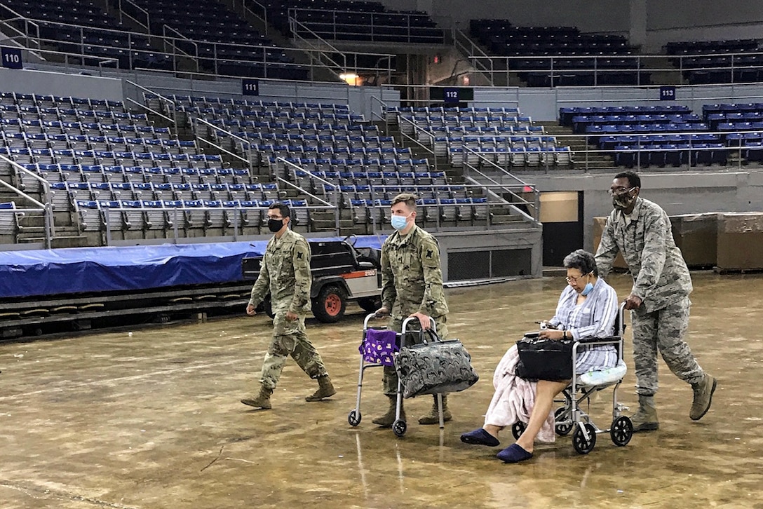 A National Guardsman pushes a citizen in a wheelchair inside an arena as two others walk beside them, one holding luggage and a walker.