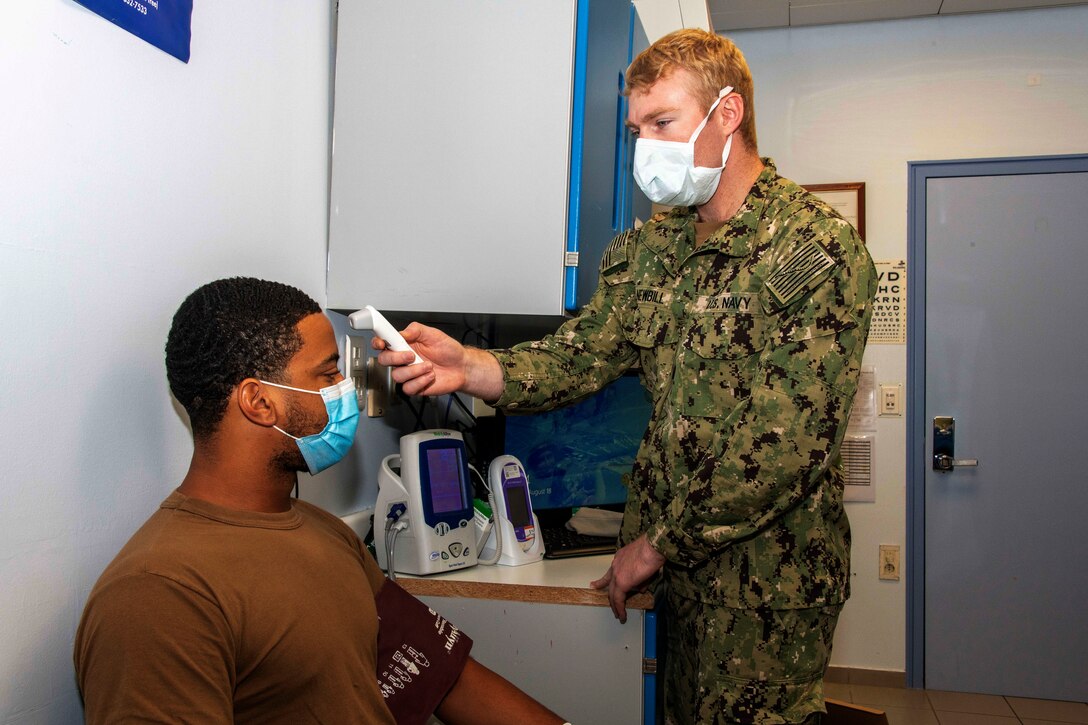 A sailor wearing a face mask takes the temperature of another sailor wearing a face mask.
