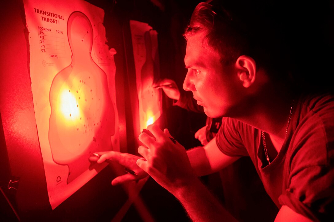 A sailor holds a flashlight and looks at a target sheet illuminated by a red light.