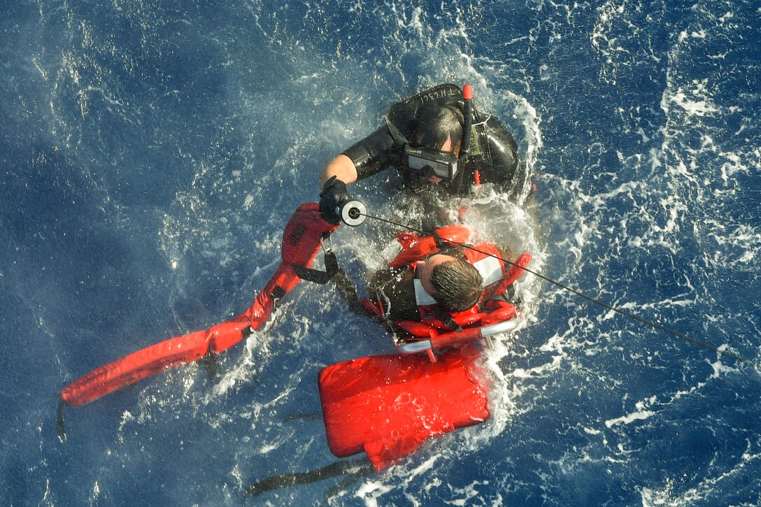 A sailor in a wet suit holds another sailor on life jacket during rescue training in a body of water.