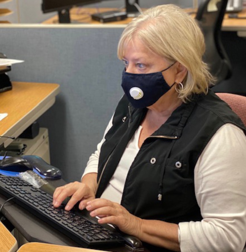 Woman wearing a face mask sits at a desk with her hands on a computer keyboard.