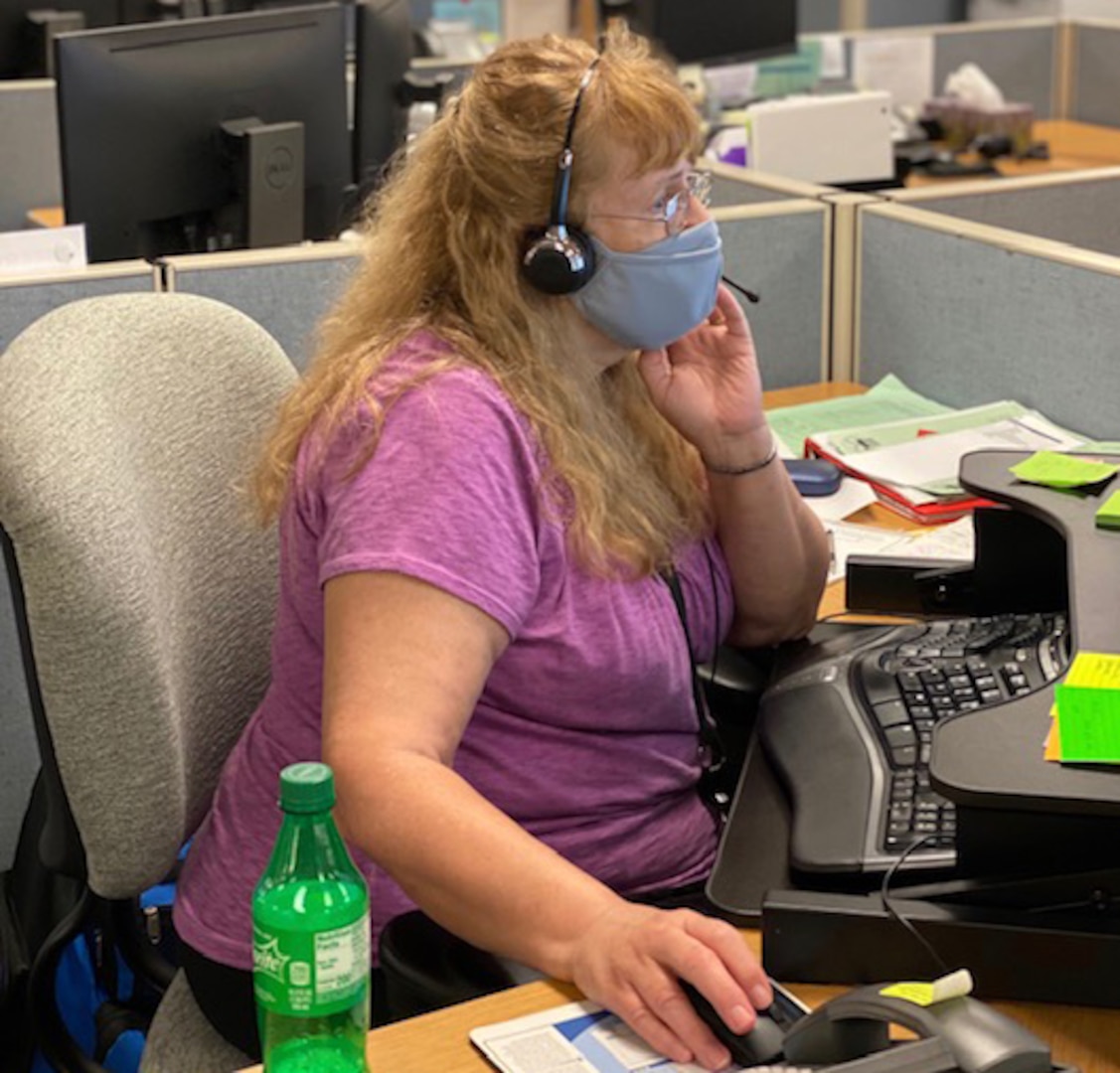 Woman wearing a face mask and headphones sits at a desk in front of a computer.