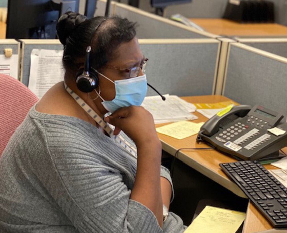 Woman wearing a face mask and headphones sits at a desk in front of a computer with a telephone nearby.