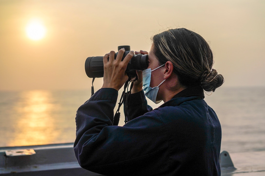 A sailor aboard a ship looks through binoculars at the open sea, with an orange sky and muted sun in the distance.