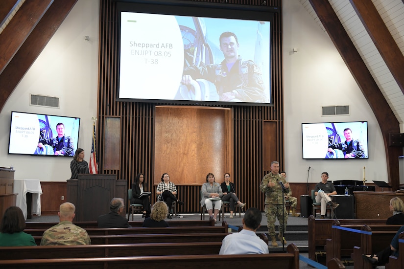 Photo shows Col. Moore standing at mic stand addressing audience with large screens behind him showing Capt. Nicholas Whitlock.