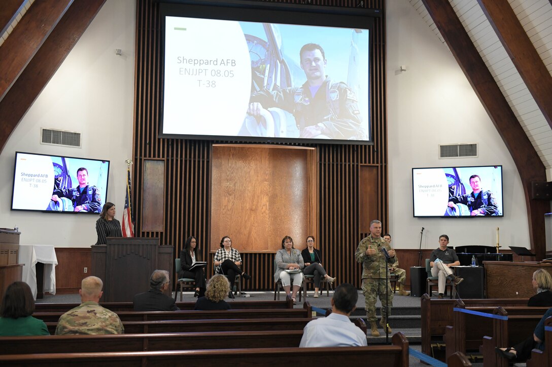 Photo shows Col. Moore standing at mic stand addressing audience with large screens behind him showing Capt. Nicholas Whitlock.