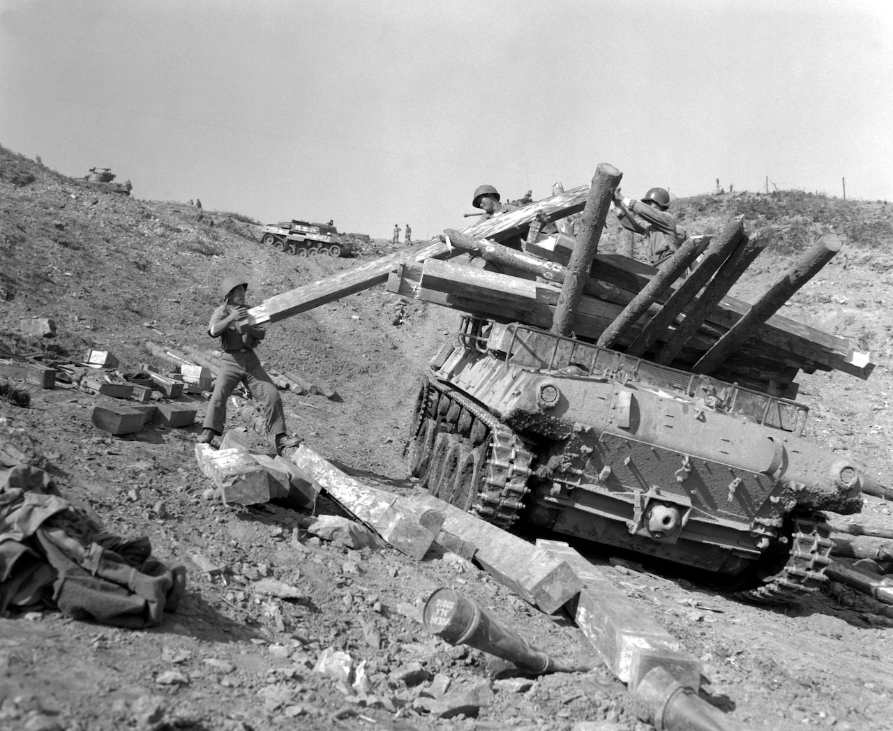 Three men load a large log onto a tank along a barren hill in a black and white photo.