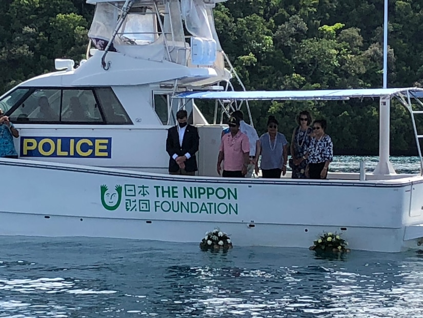 Five people in a boat look at wreaths floating on the water. Other people are in the background.