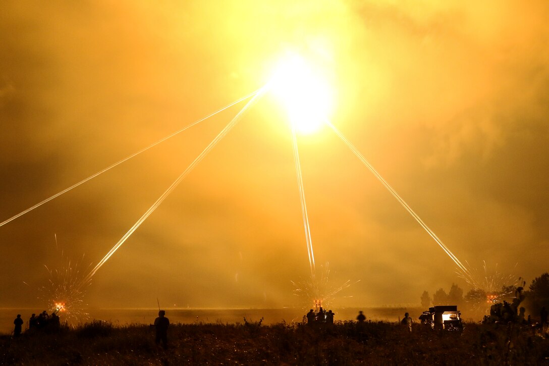 Service members in silhouette on the ground fire weapons that light up a yellowish sky.