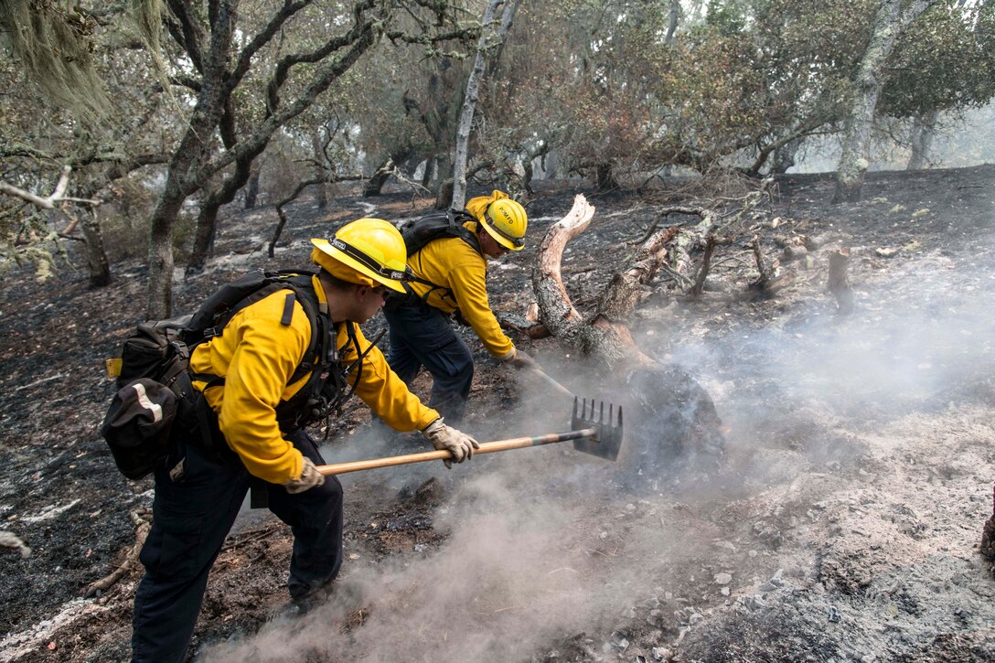 Two Army firefighters use tools on smoldering ground.