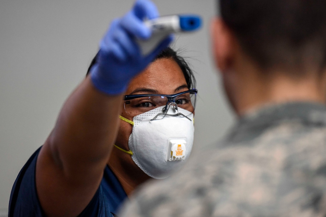 A woman wearing a face mask hold a thermometer up to the forehead of a aims a military service member.
