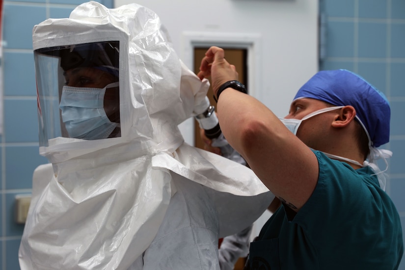 A medical technician wearing a face mask helps someone put on personal protective equipment.