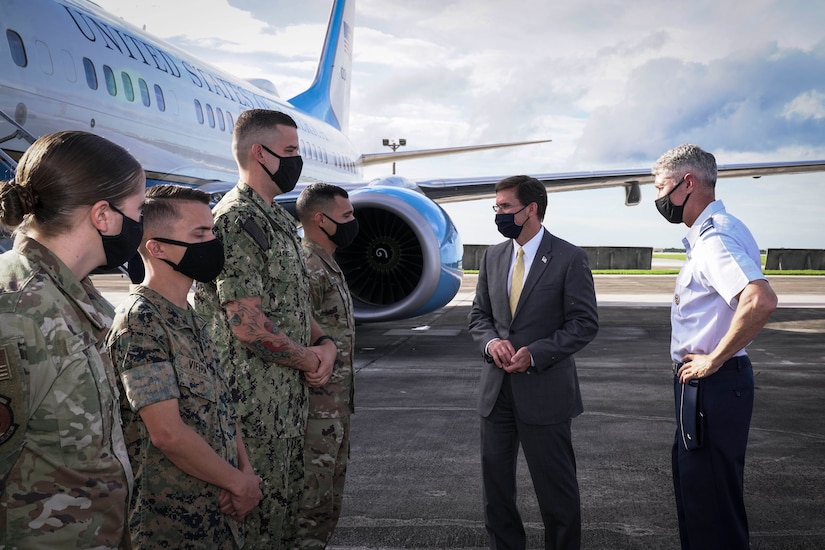 Man in civilian business suit greets military personnel on the tarmac. All are wearing masks. Official airplane is in the background.