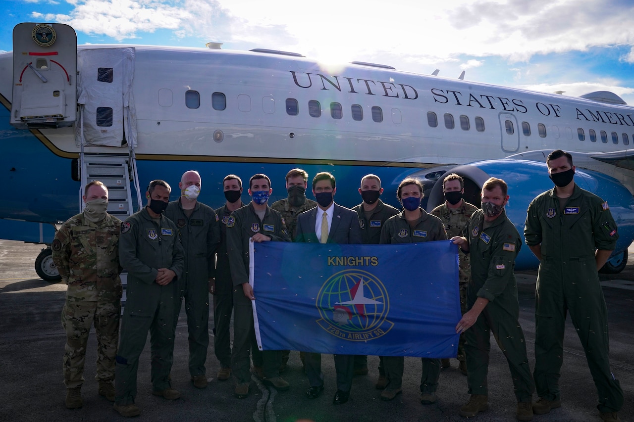 A man in business attire stands with men in military uniforms. All are wearing masks. An official airplane is in the background.