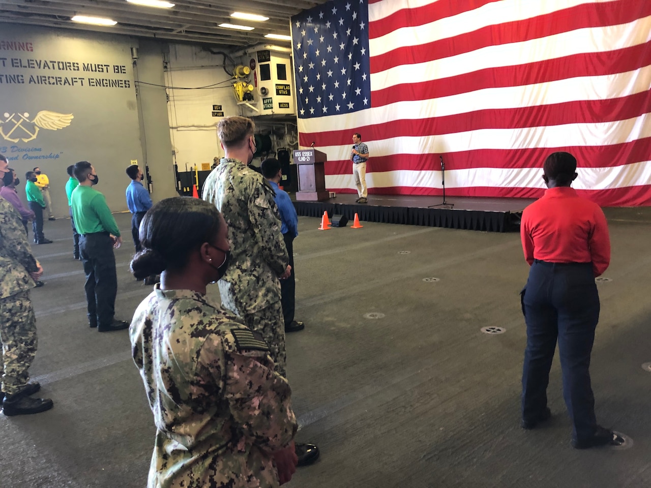 A man standing in front of a large U.S. flag speaks to a group of people who are socially distanced.