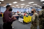 Man wearing a mask speaking to a woman in military uniform. People sewing clothing in the background.