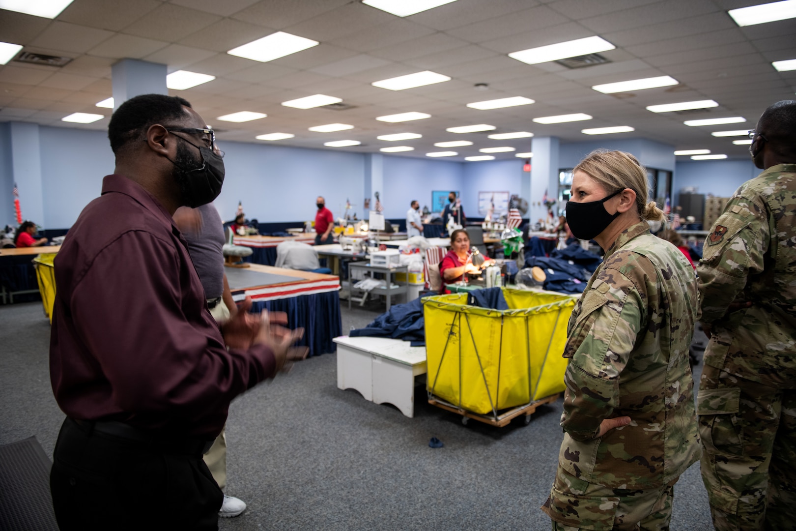 Man wearing a mask speaking to a woman in military uniform. People sewing clothing in the background.