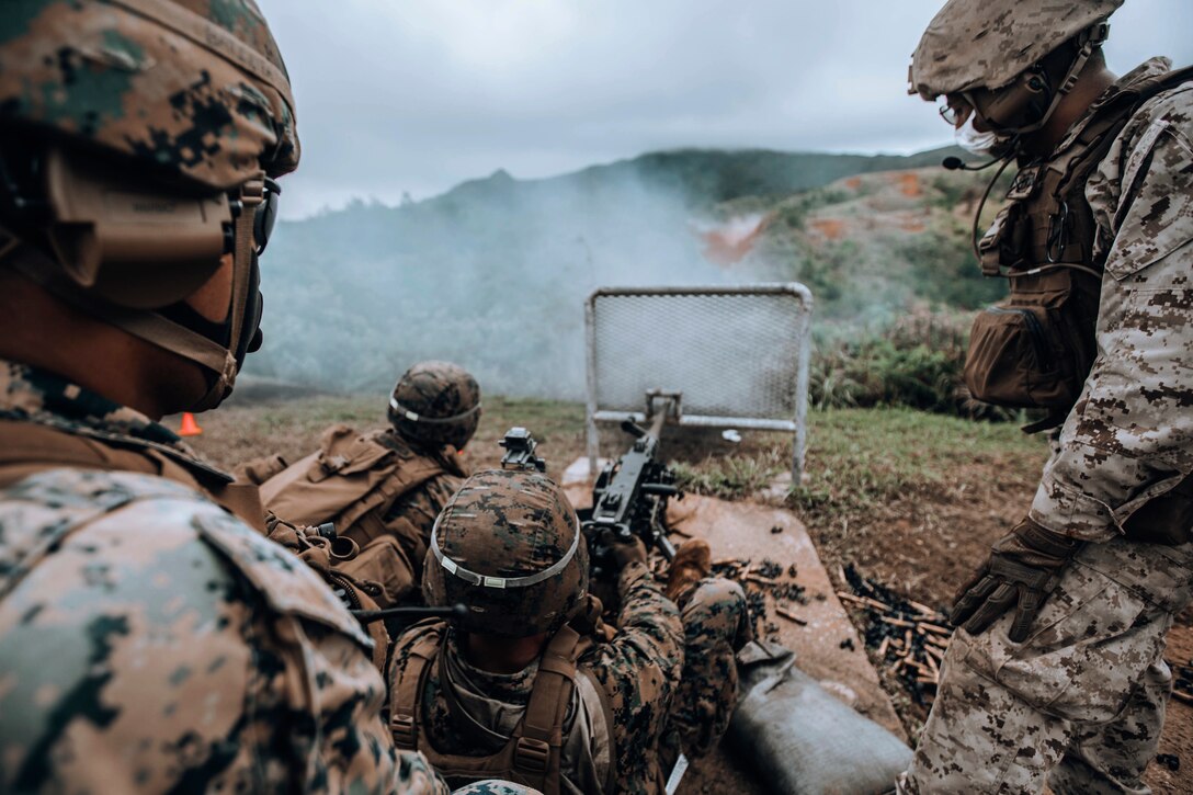 A Marine fires a weapon in a field-like area as other Marines watch.