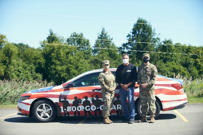 Two people in military uniforms and a civilian stand in front of a car with “1-800-Go-Guard” painted on the side.