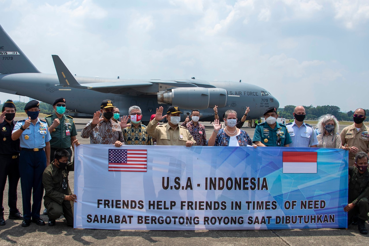 With a large plane in the background, a group of more than 20 people pose for a photo while holding a banner with the words “USA-Indonesia Friends Help Friends in Time of Need.