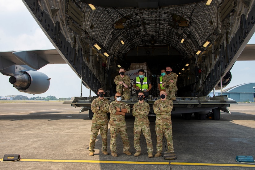 Eight service members wearing face masks stand at the back of a cargo plane and pose for a photograph.