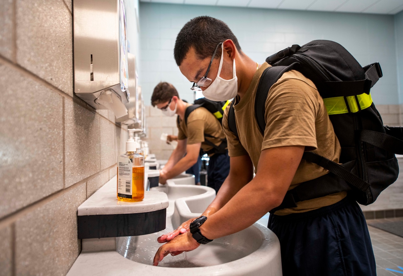 Navy recruits wearing backpacks and face masks wash their hands in bathroom sinks.