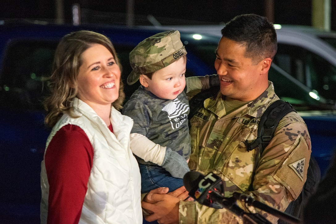 A soldier holds his child while his wife smiles.