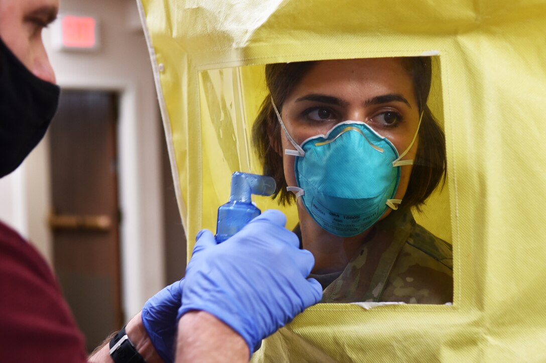 A woman wearing protective gear and an N95 mask looks to her left while a person wearing surgical gloves points a bottle at her face.