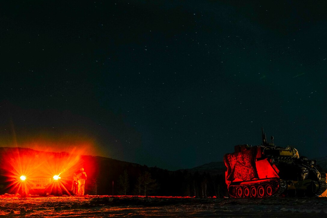 A parked military vehicle shines red lights at night next to another vehicle.