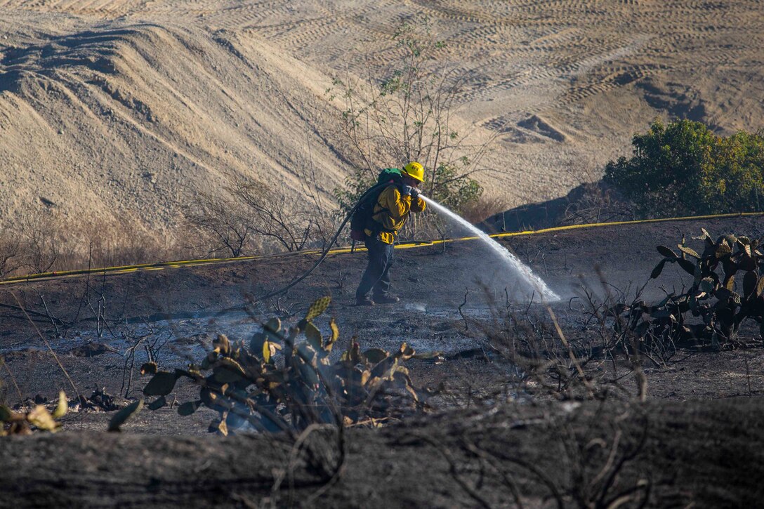 A firefighter wearing fire protection gear uses a hose to spray water on the ground.