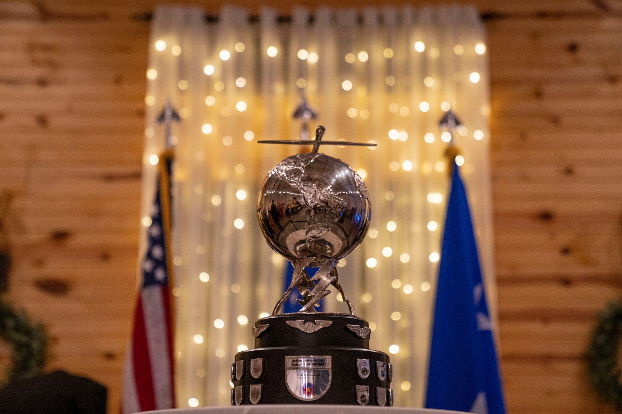 The rotating General Atomics Remotely Piloted Aircraft Squadron of the year trophy which involves a silver metal globe propped up by twisted, metal lightning bolts and topped with a metal RPA sitting in front of the United States flag, the U.S. Air Force flag and white holiday lights.