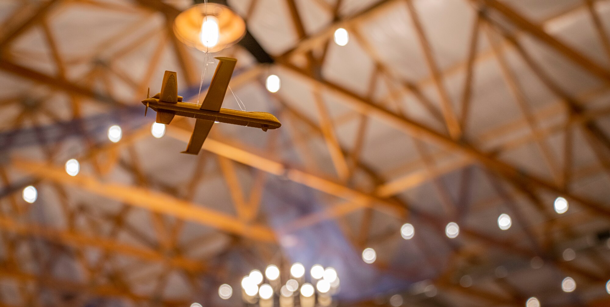 A small, wooden MQ-9 Reaper figurine hangs on a string with white holiday lights and wooden ceiling structures in the background.