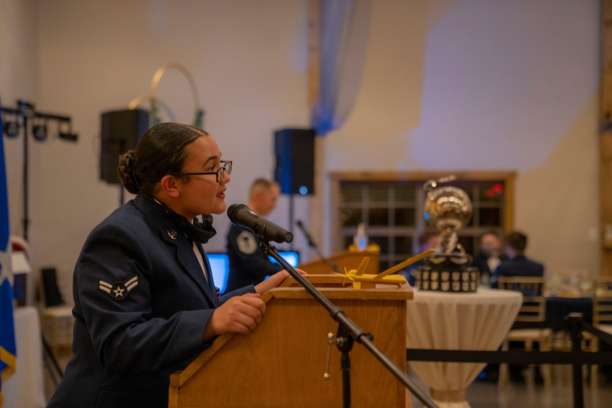 A female Airman leans into a podium to speak to the audience while a male Airman speaks at another podium in the background.