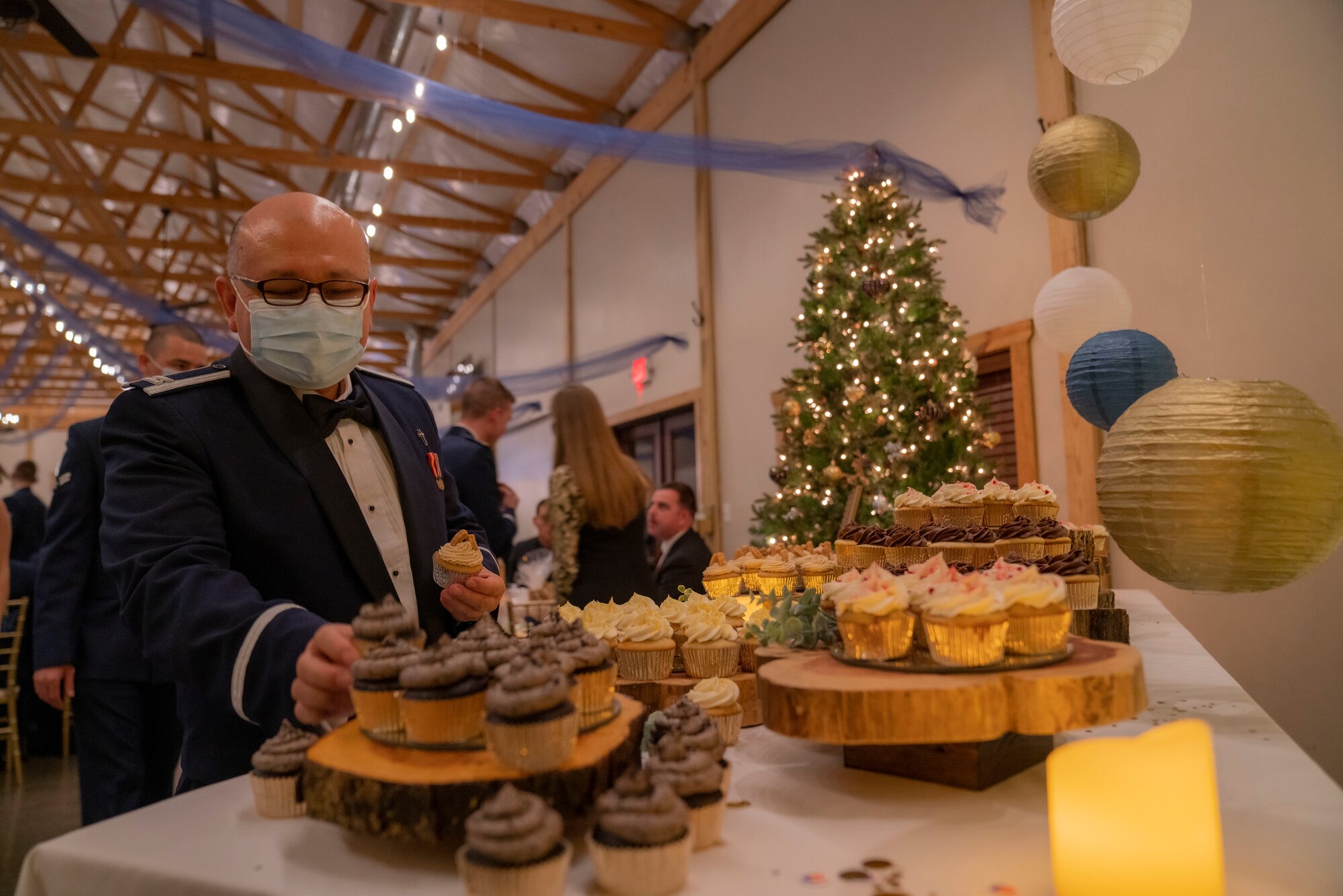 A male Airman is grabbing a cupcake from a cupcake stand with a Christmas tree in the background.