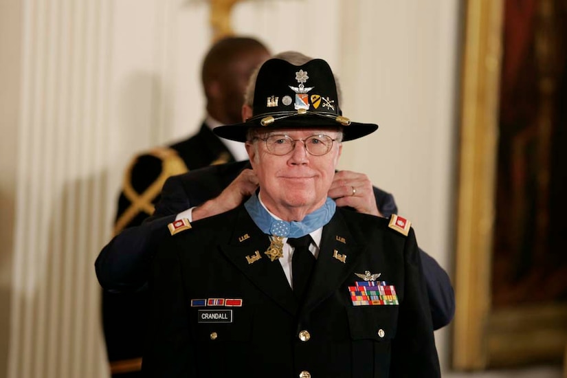 A man wearing a brimmed hat stands while a second man puts a medal around his neck.