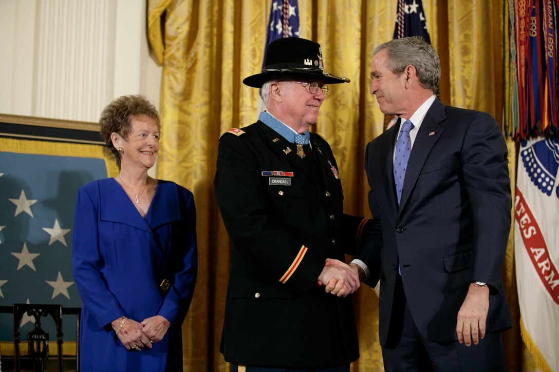 Two men shake hands while a woman in the background watches.