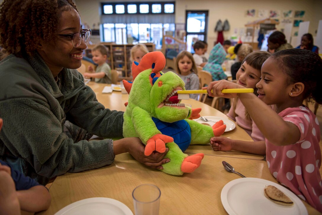 A smiling child brushes a stuffed animal's teeth as an airman and other children watch and smile.