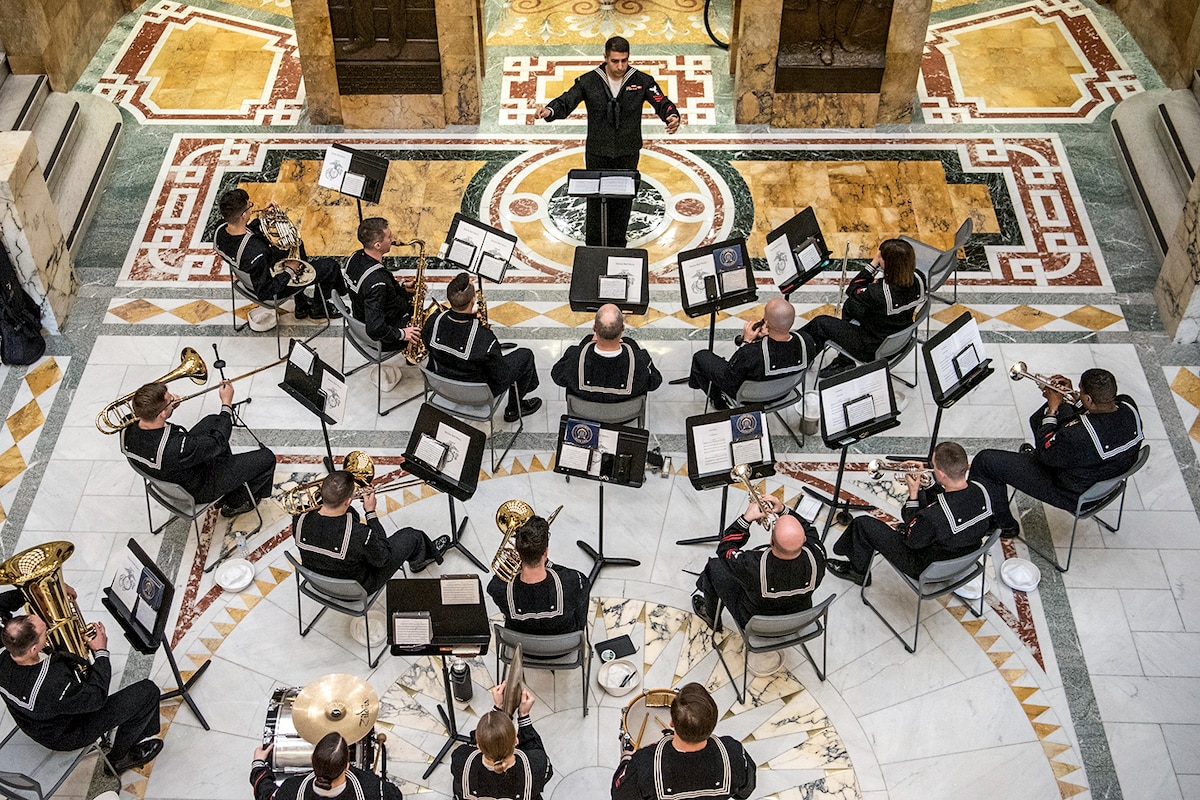 A Navy conductor leads a group of Navy musicians, shown from overhead in an ornate room.