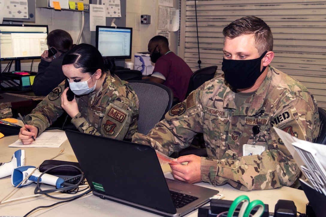 Two airmen wearing face masks use a computer and phone to contact patients prior to testing.
