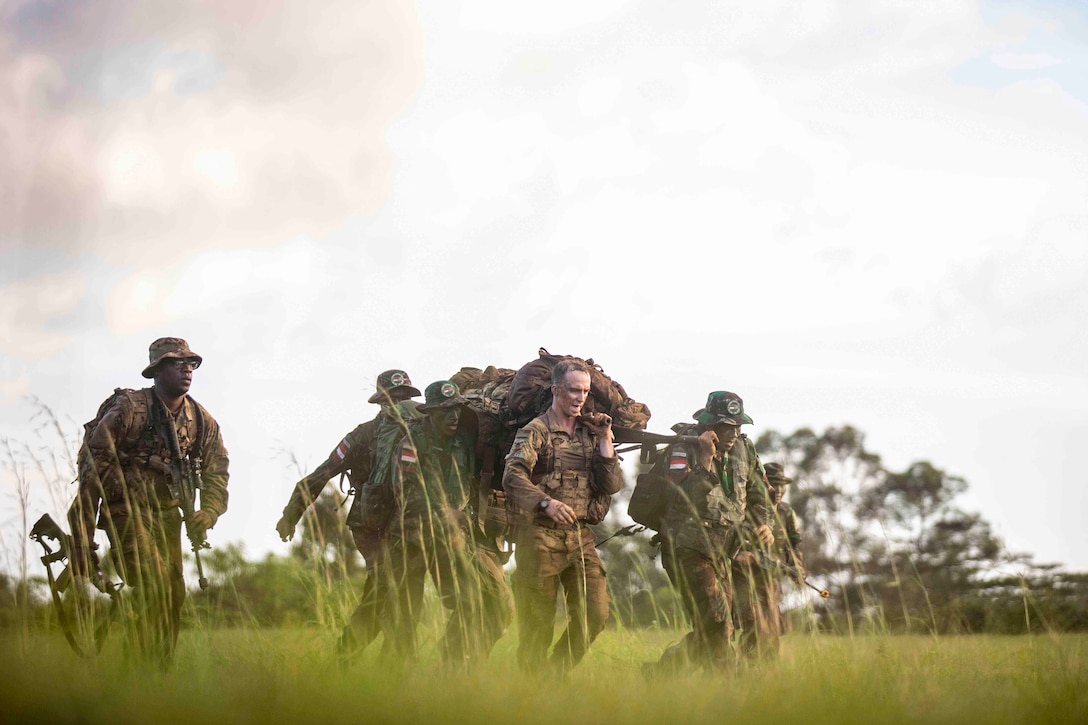 A group of soldiers walk through a grassy field carrying a stretcher.