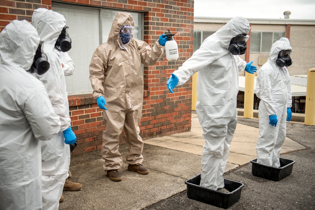 A civilian in protective gear sprays a service member in protective gear standing in bins outside a building.