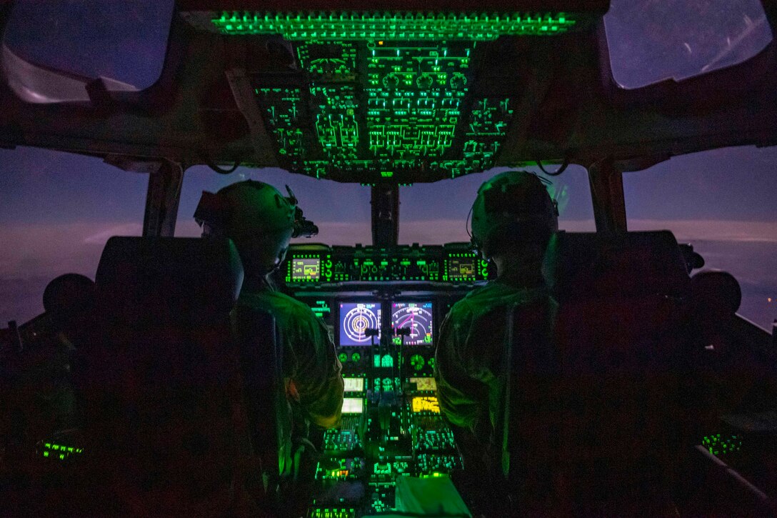 Two pilots sit in a cockpit illuminated by green light.
