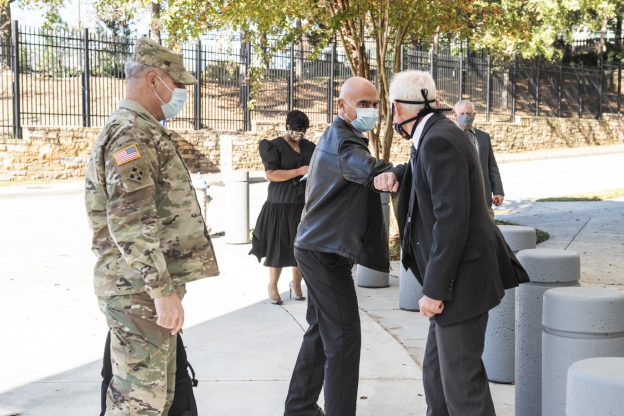 Two men bump elbows as a man in a military uniform watches.