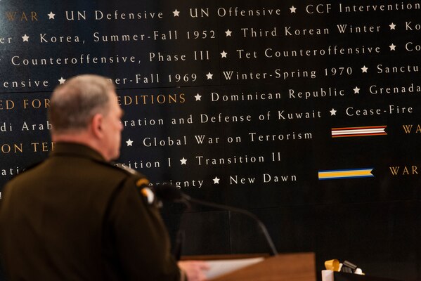 A man is seen from behind as he stands behind a lectern; he faces large words written on a wall.