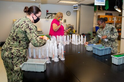 Volunteer Sailors Third Class Petty Officer Sarah Wise and Second Class Petty Officer Khadijah Sam help put together science experiment packets the teachers are sending to the students at home so they can complete the assignment at home self-sufficiently.