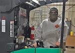 A masked man drives material handling equipment in a warehouse.