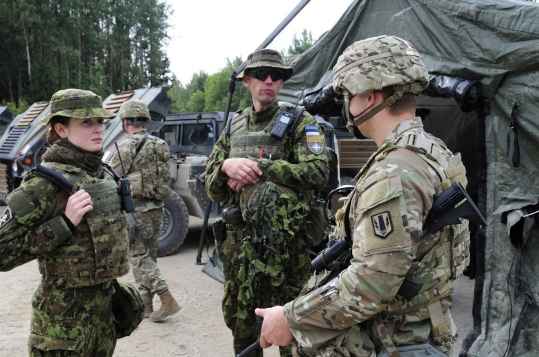 Three soldiers in combat uniforms talk outside a tent.
