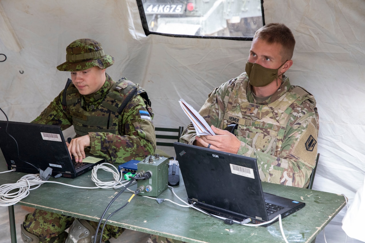 Two soldiers in combat uniforms sit behind laptops at a table.