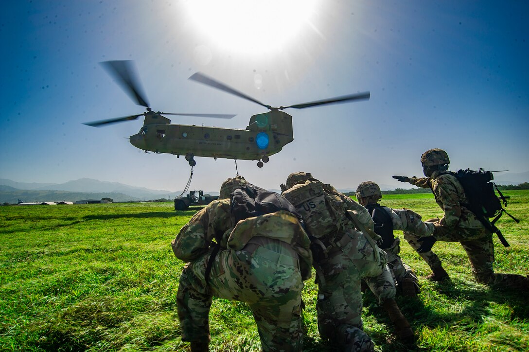 Soldiers wait to attach a sling to a descending helicopter.